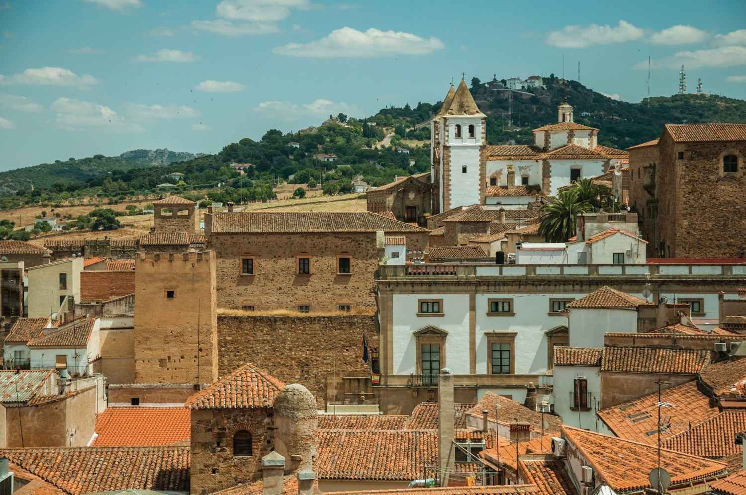 Vista de una ciudad histórica con techos de tejas rojas, edificios de piedra y una torre de iglesia bajo un cielo azul.
