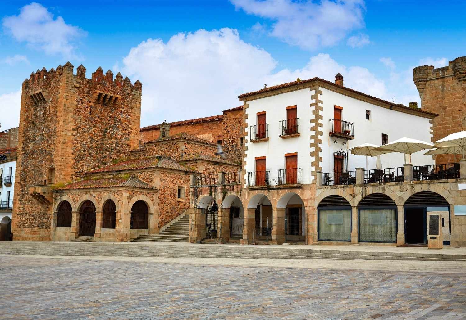 Castillo de piedra y edificios blancos con fachadas arqueadas en una plaza, bajo un cielo azul.