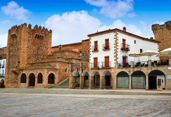 Castillo de piedra y edificios blancos con fachadas arqueadas en una plaza, bajo un cielo azul.