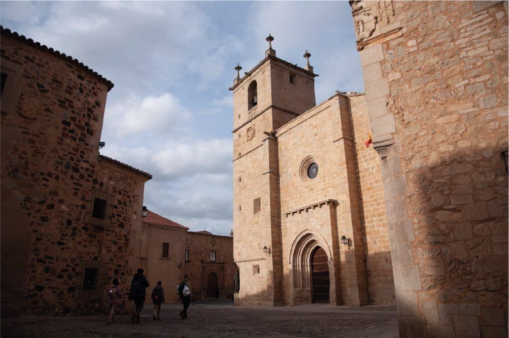 Iglesia de piedra con puerta arqueada y campanario en una plaza adoquinada, con algunas personas caminando.