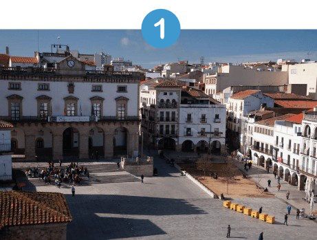 Vista de una plaza de la ciudad con edificios históricos y una torre de reloj bajo un cielo brillante.