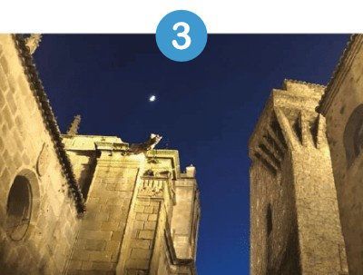 Edificios de piedra gótica recortados contra un cielo azul oscuro vespertino con una luna creciente.