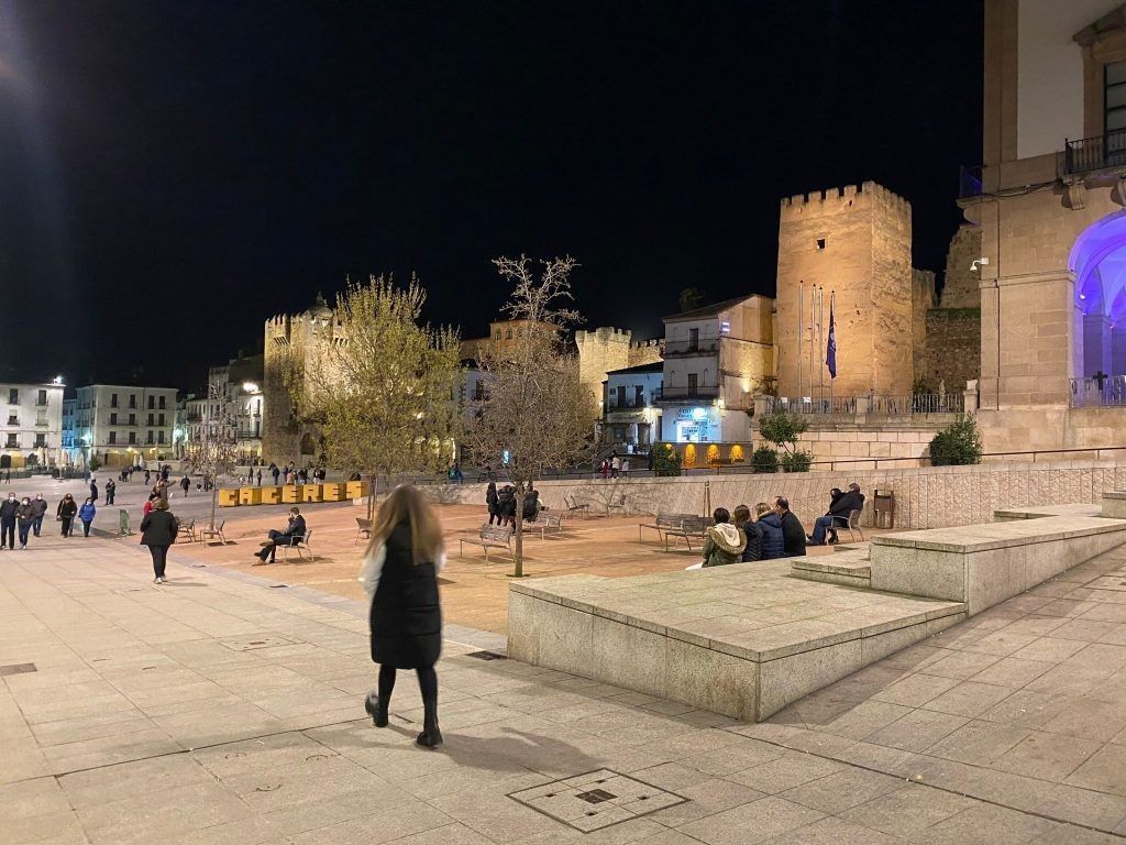 Vista nocturna de una plaza de la ciudad con un castillo al fondo, gente caminando y sentada en los bancos.