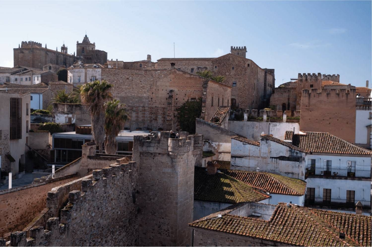 Paisaje medieval con edificios de piedra, techos de tejas y un castillo en la cima de una colina bajo un cielo azul.
