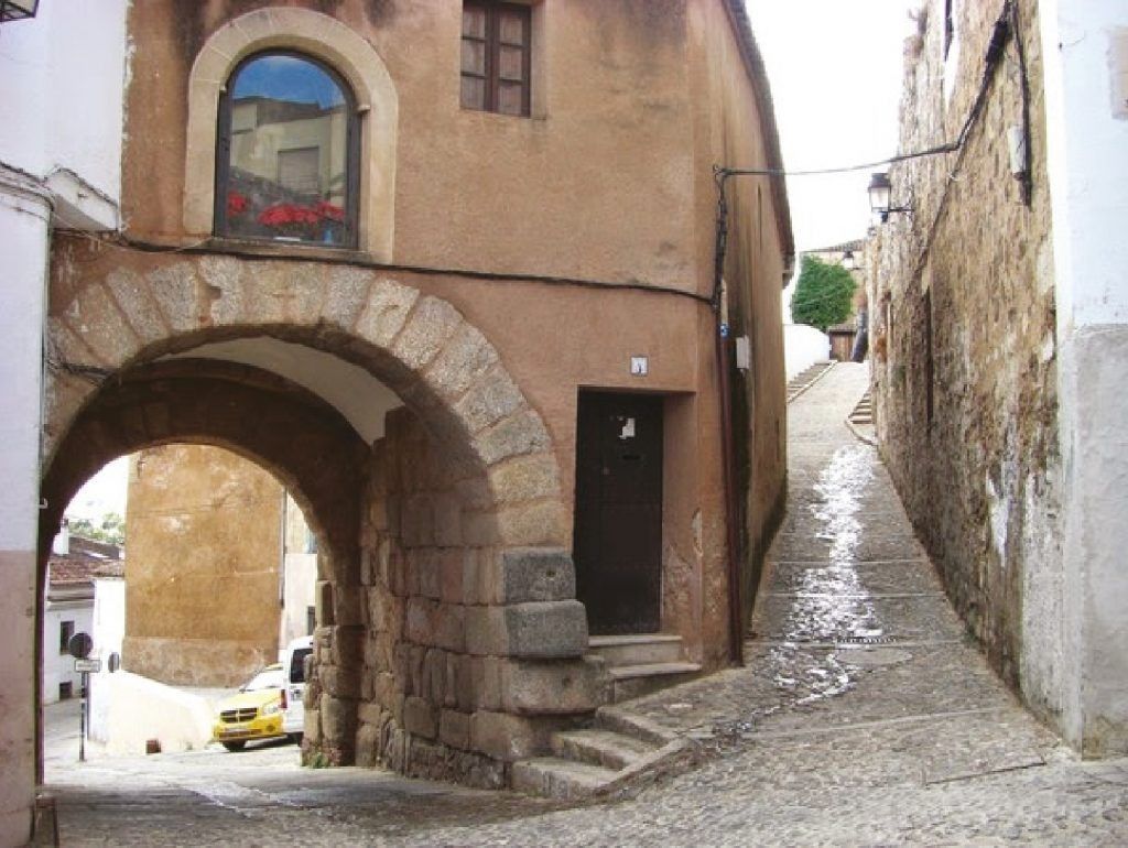 Un arco de piedra conduce a una calle estrecha y en pendiente de una ciudad; edificios marrones y una ventana con flores.