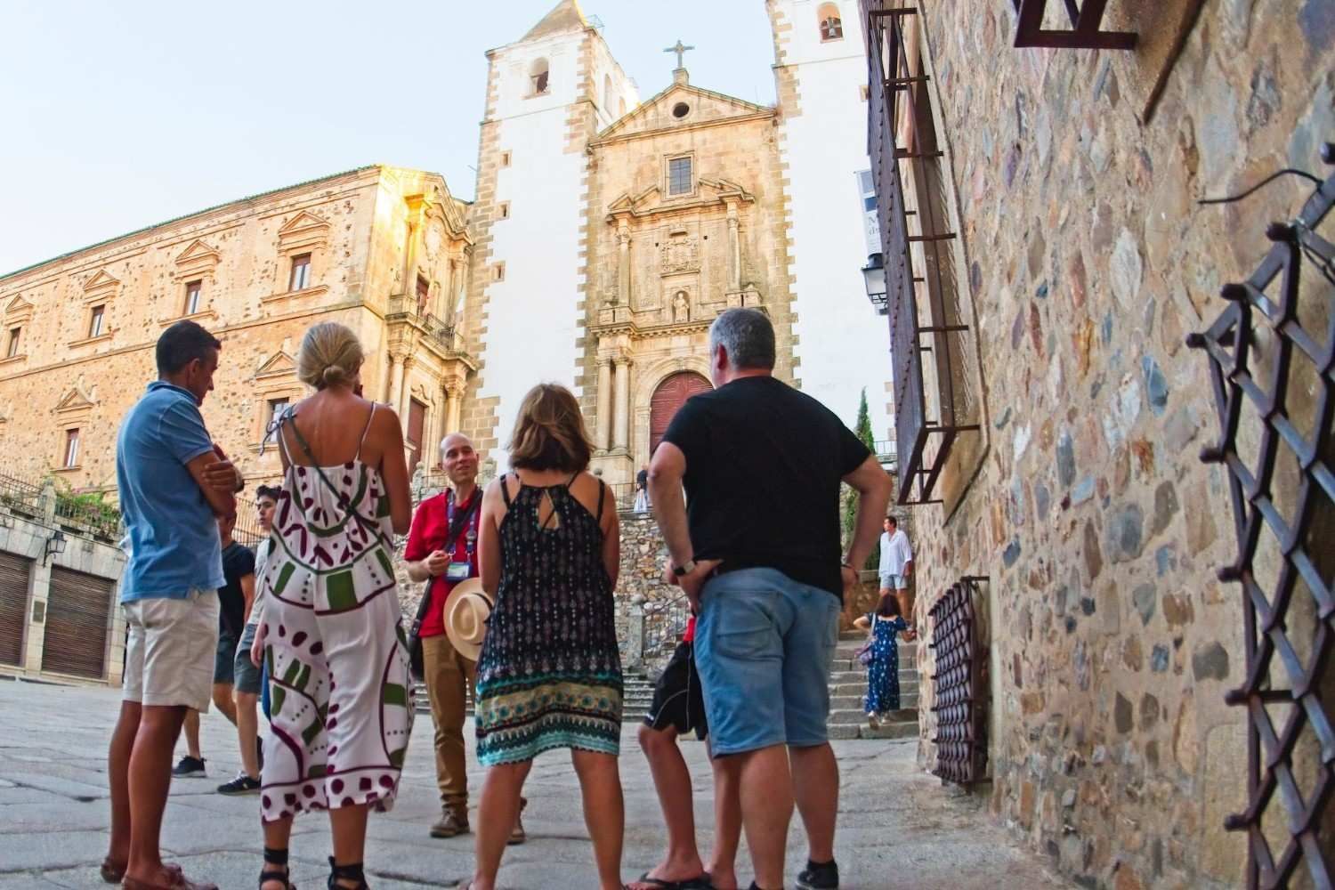 Turistas contemplando una iglesia histórica, España. Vista exterior: edificios de piedra, luz cálida.