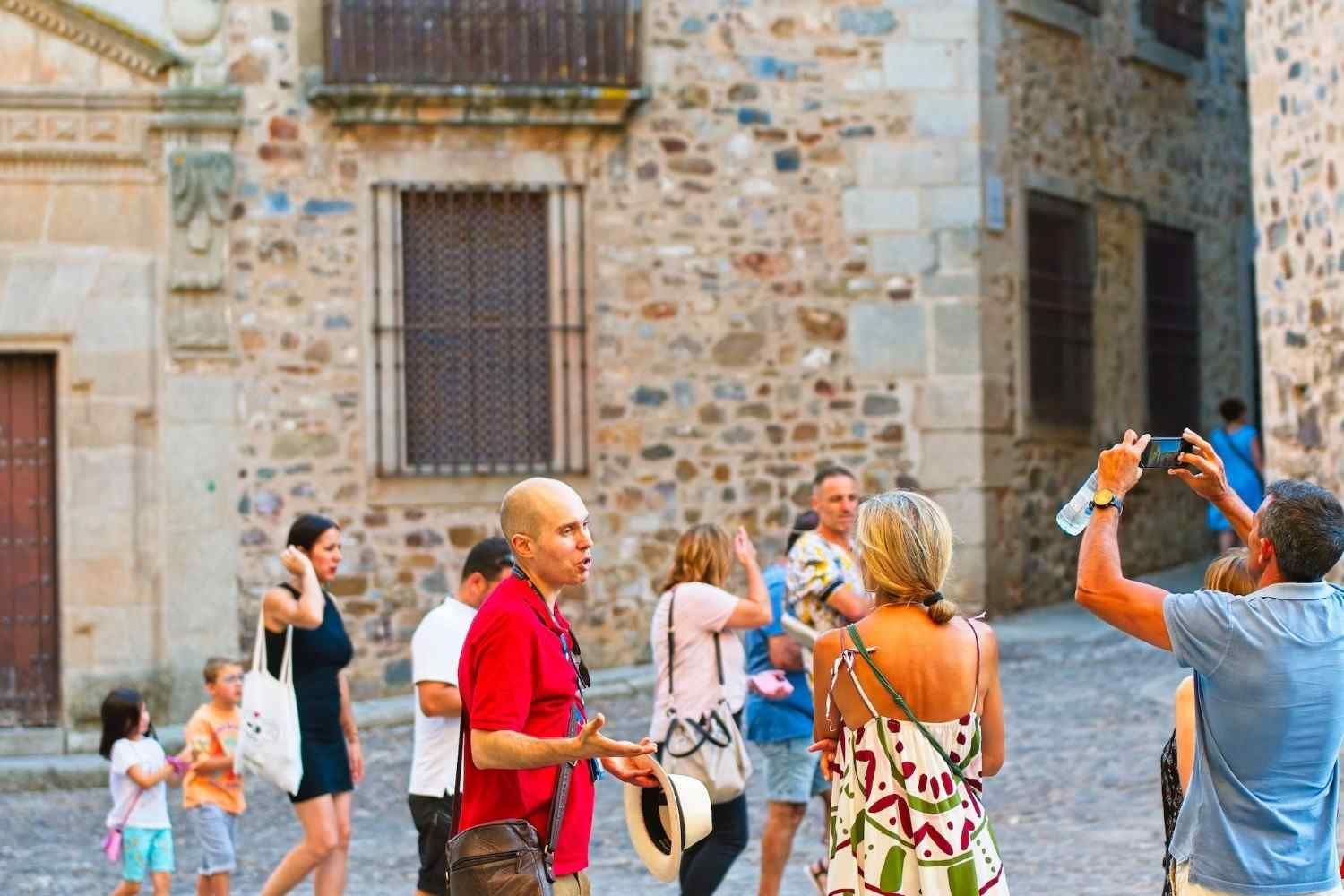 Gente caminando por una calle empedrada, algunos mirando edificios, uno tomando una foto. Escena soleada al aire libre.
