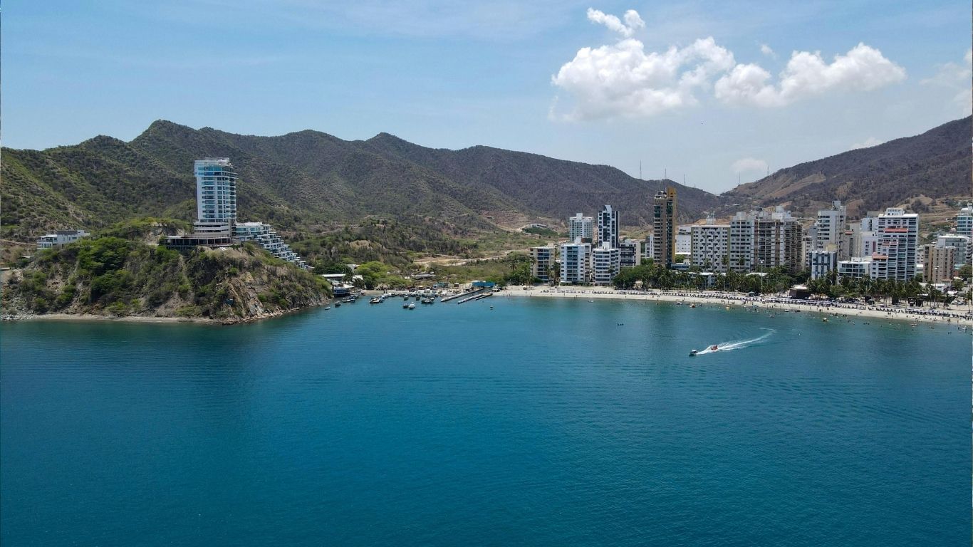 Coastal cityscape with mountains, buildings, and boats on a clear, blue ocean under a sunny sky.
