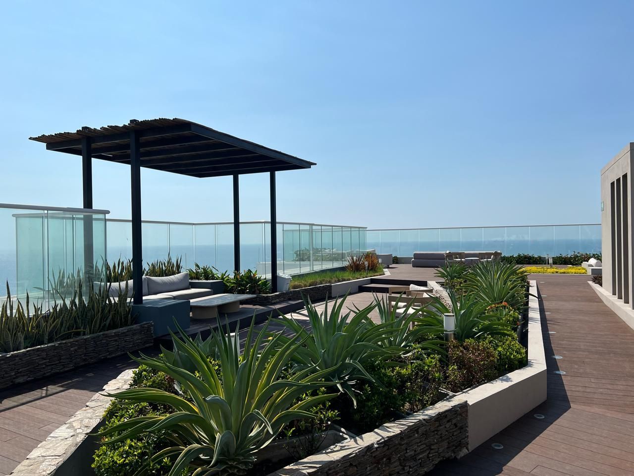 Rooftop garden with ocean view, featuring a pergola, plants, seating, and glass barriers on a sunny day.
