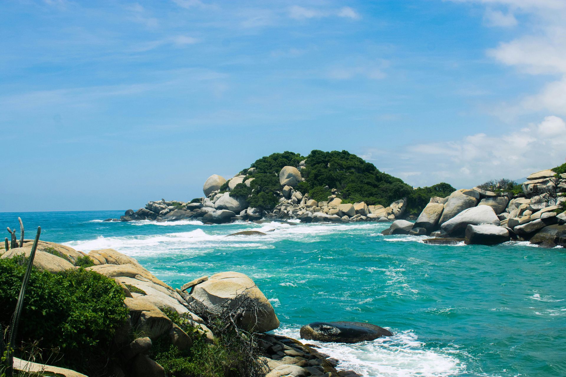 Turquoise ocean waves crashing against rocky shoreline under a blue sky, island in the background.