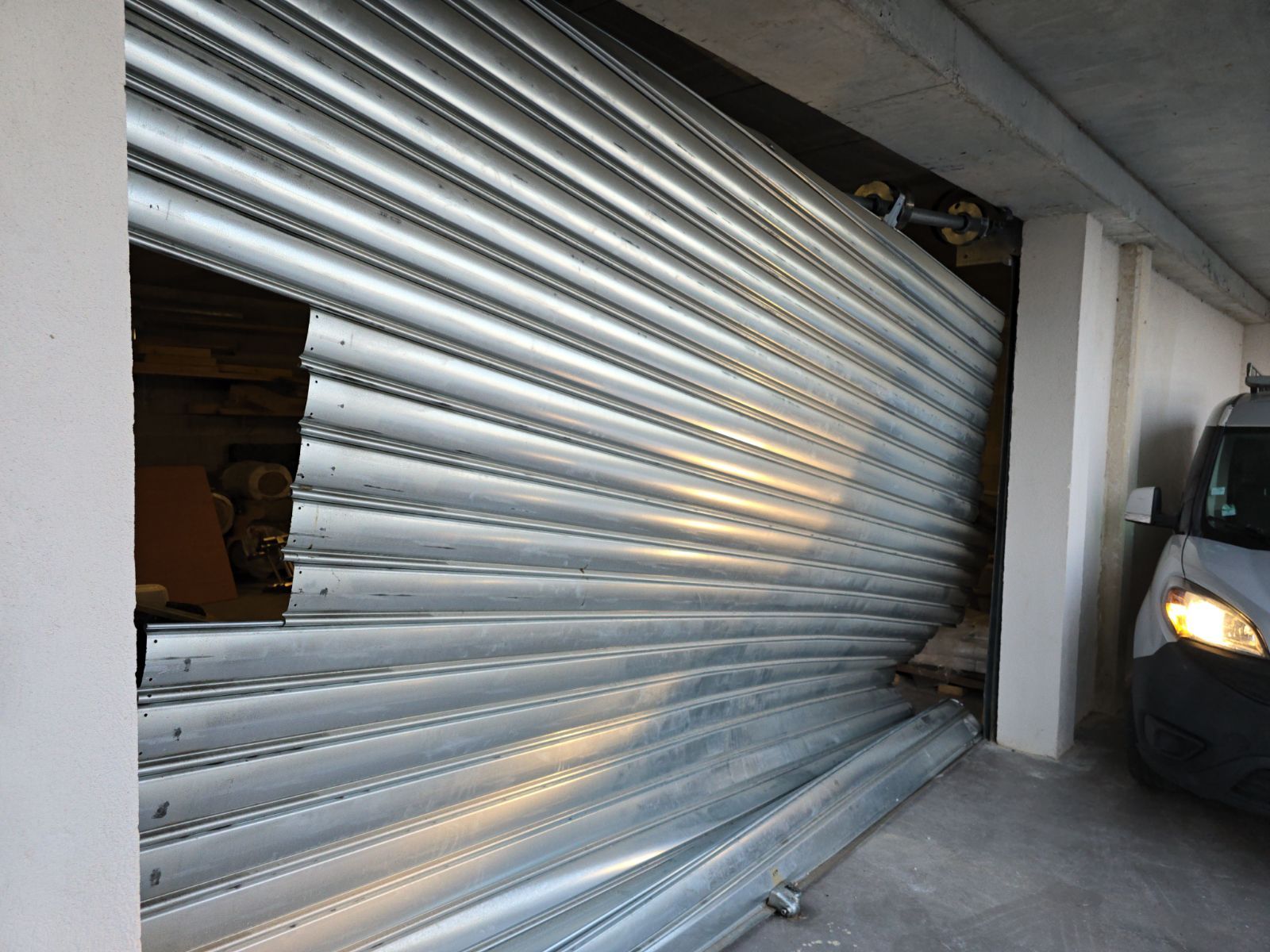 Porte de garage sectionnelle en métal argenté partiellement fermée, avec un véhicule noir à côté.