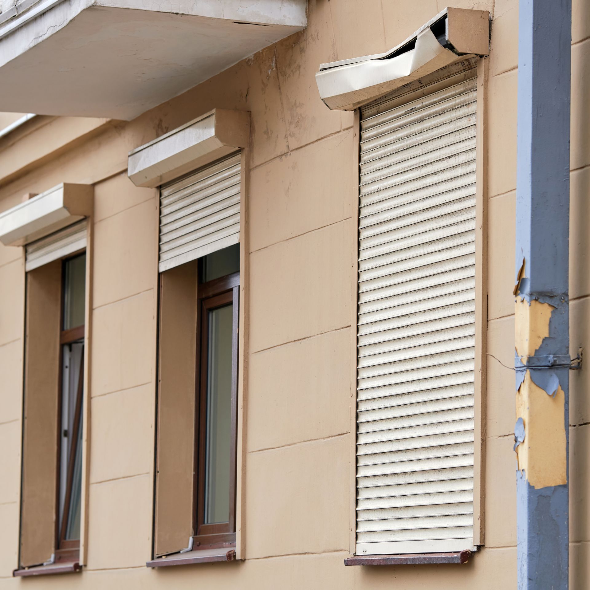 Façade d'immeuble endommagée avec trois fenêtres et volets roulants blancs, peinture écaillée sur le bord droit