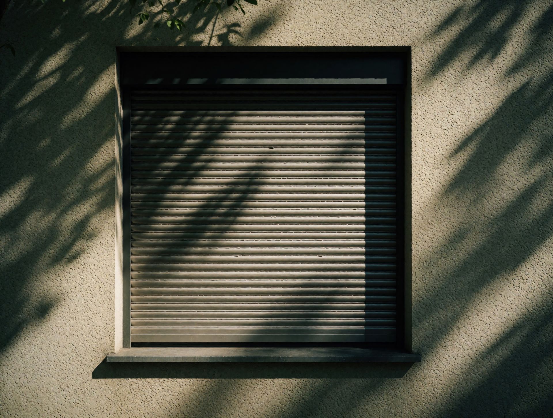 Fenêtre fermée avec stores horizontaux sur un mur beige, ombragée par des arbres.