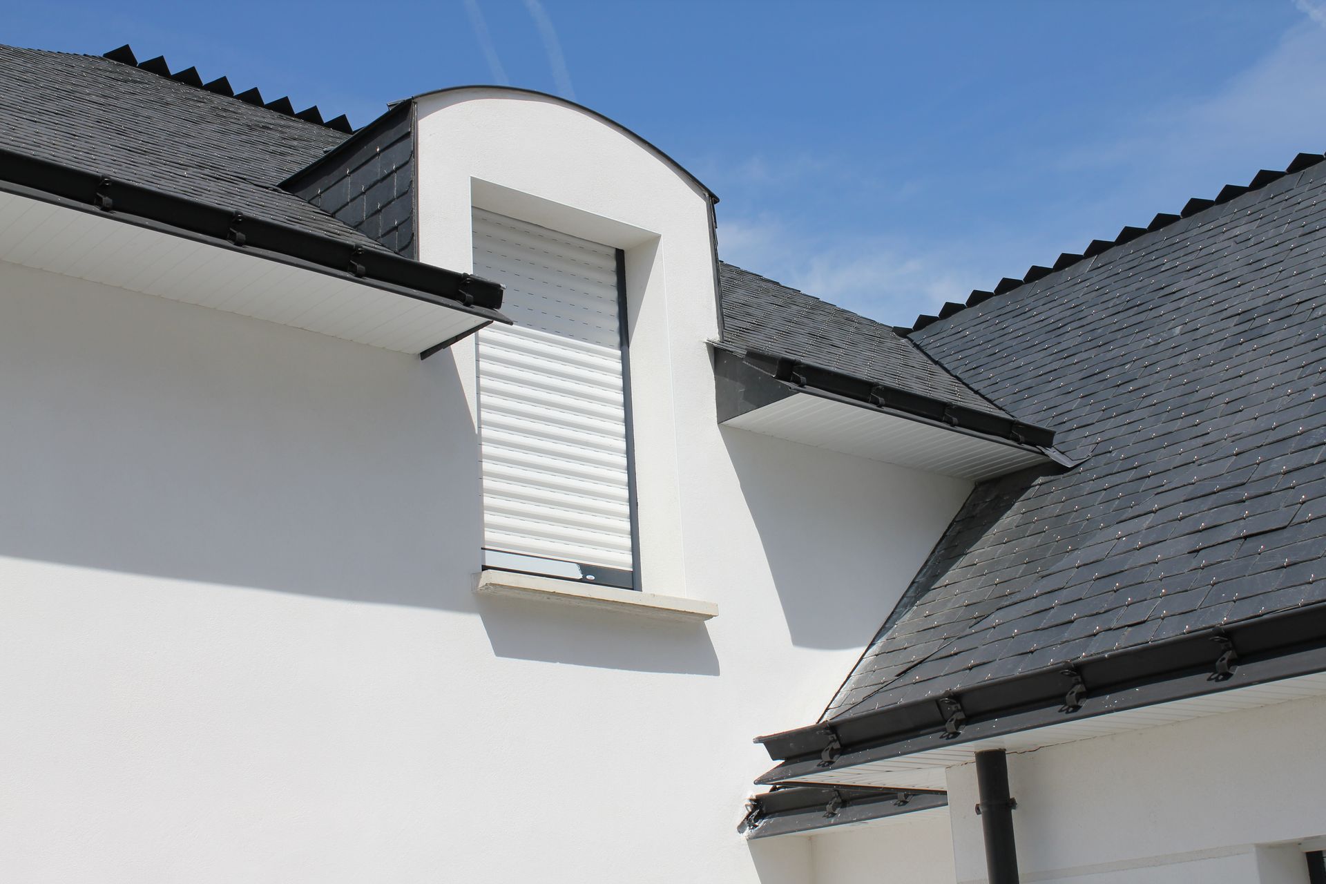 Extérieur d'une maison blanche avec un toit gris foncé et une petite lucarne sous un ciel bleu