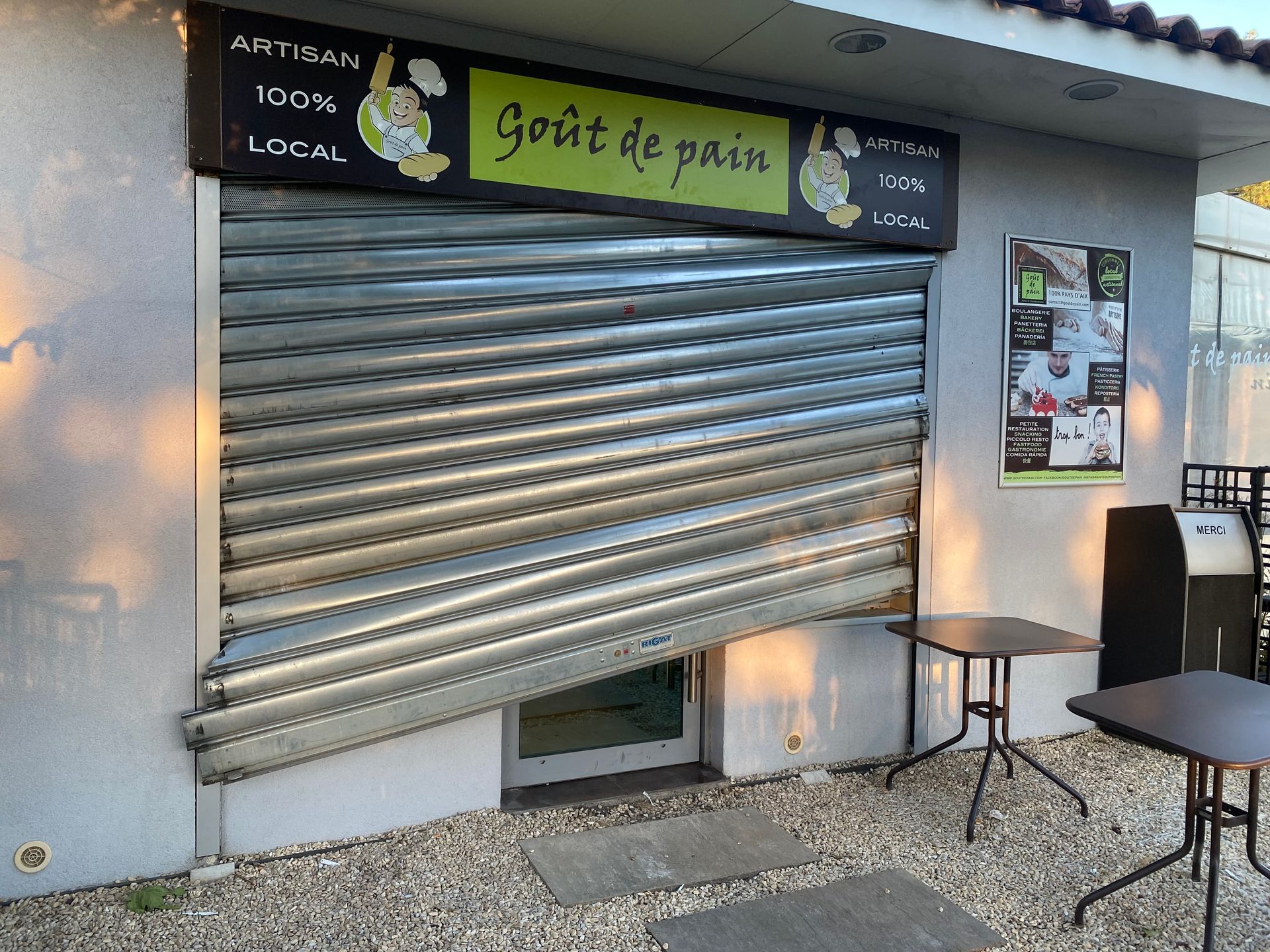 Façade fermée avec volet métallique, enseigne et tables en terrasse sur le trottoir.