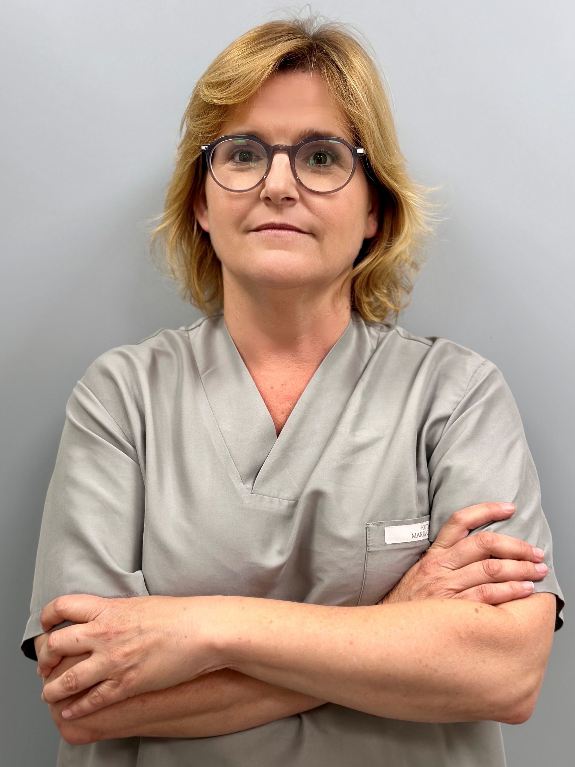 Una mujer que lleva gafas y uniforme médico está de pie con los brazos cruzados.