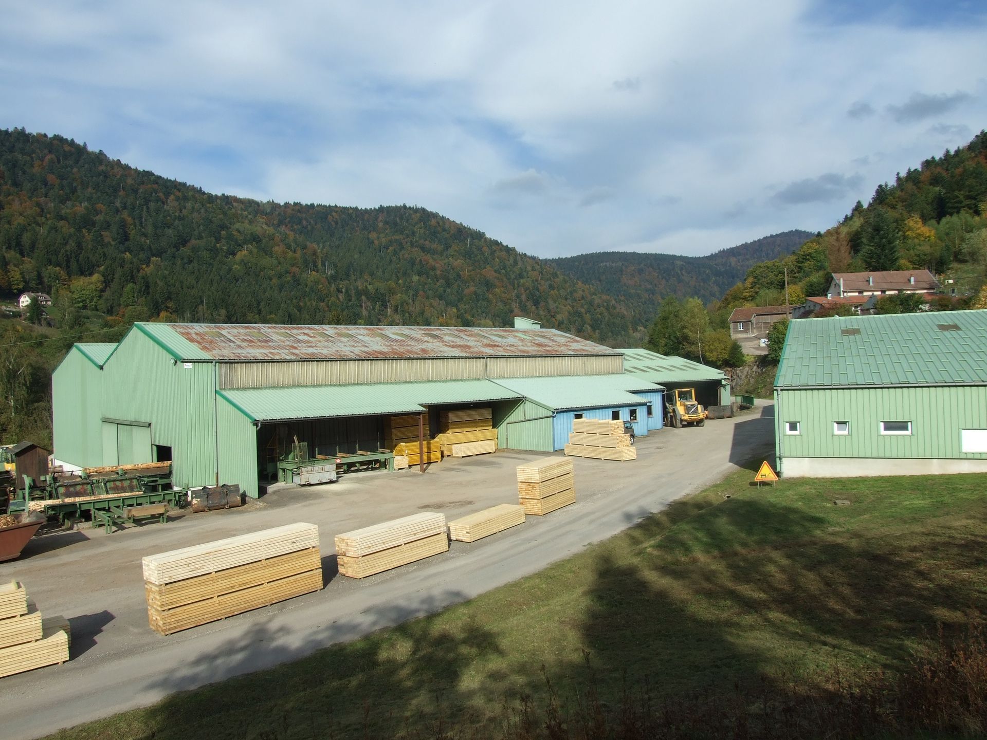 Scierie avec de longs hangars verts, des planches empilées et des collines boisées sous un ciel nuageux