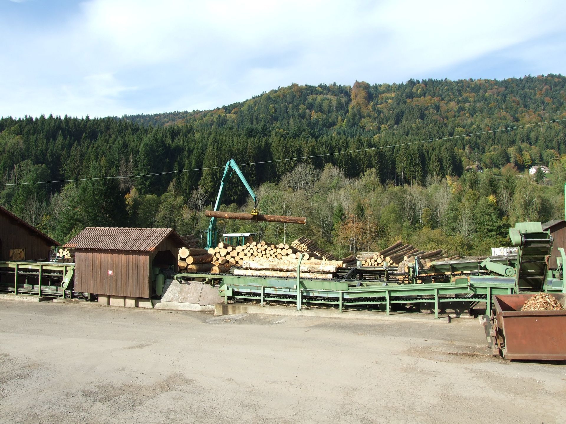 Parc à grumes avec des grumes empilées, une grue verte et des collines boisées en arrière-plan.