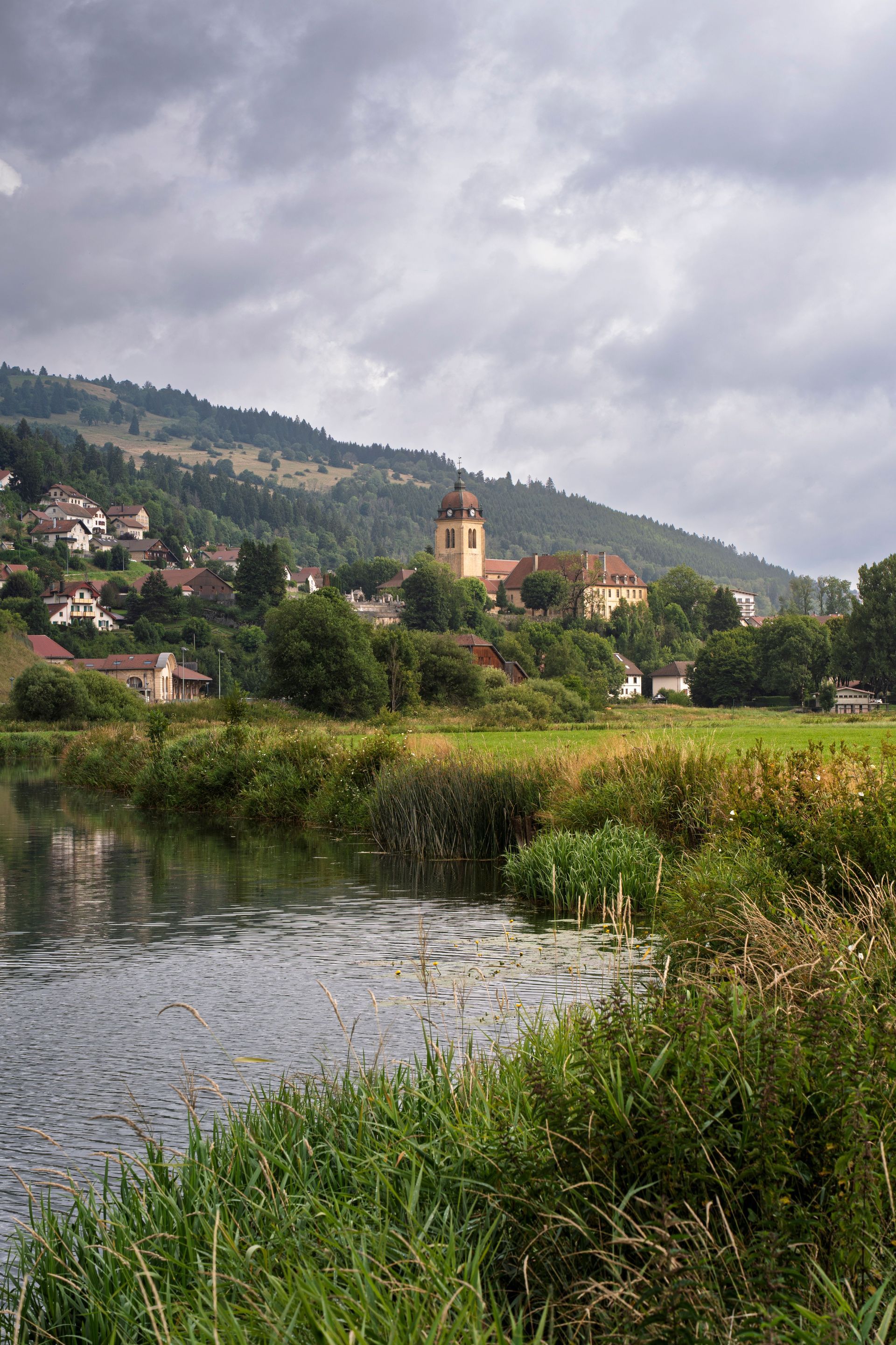 Église au bord d'une rivière, avec des berges herbeuses et des collines sous un ciel nuageux