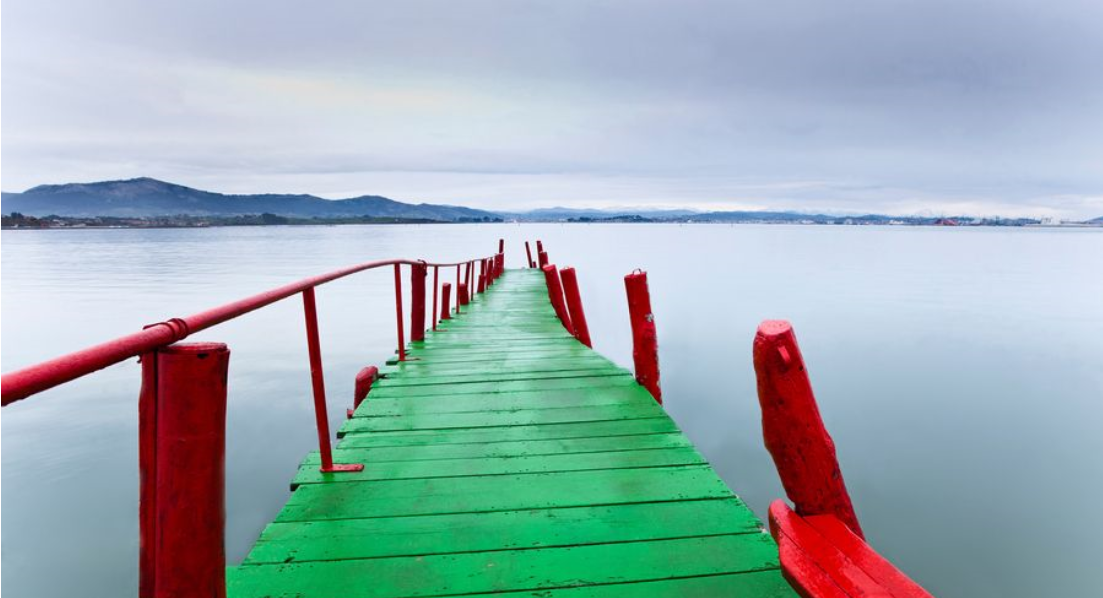 Muelle de madera roja con tablones verdes que se adentran en aguas tranquilas bajo un cielo nublado.