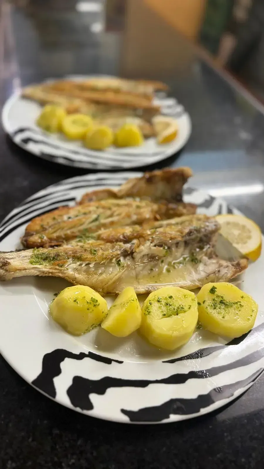 Pescado a la parrilla con patatas, servido en platos blancos y negros en la mesa de un restaurante.