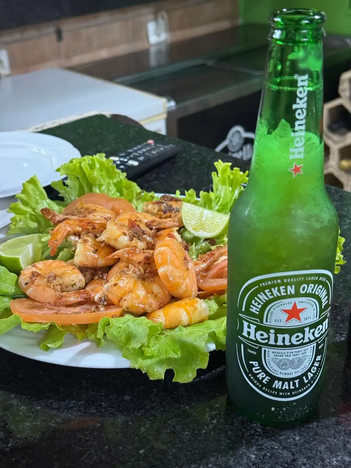 Una botella de Heineken junto a un plato de gambas glaseadas sobre hojas de lechuga en la mesa de un restaurante.