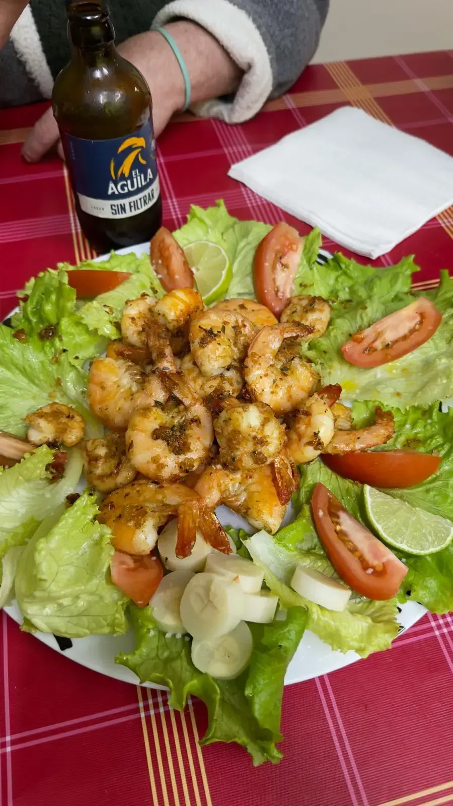 Ensalada con lechuga, tomate y camarones a la parrilla sobre un mantel a cuadros rojos, con una botella de salsa al lado.