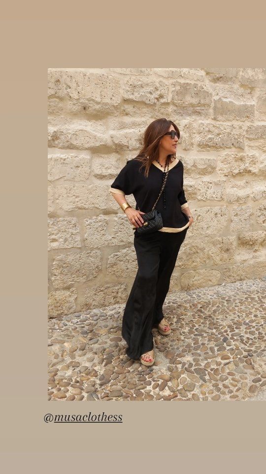 Una mujer con una camisa negra y pantalones negros está parada frente a un muro de piedra.