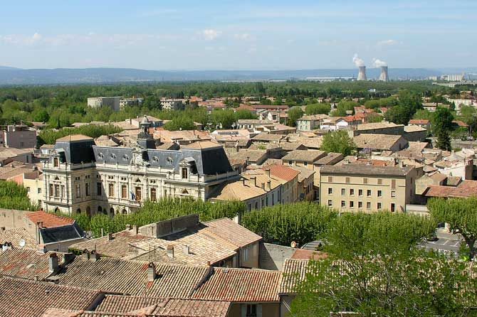 Vue des toits de tuiles en terre cuite, un grand bâtiment en pierre et des tours de refroidissement au loin.