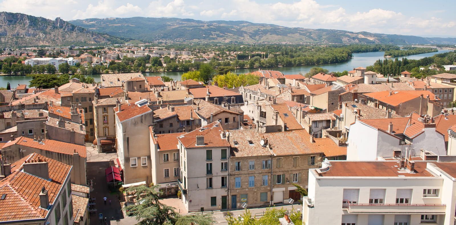 Vue d'une ville aux toits de tuiles rouges, avec une rivière et des collines ondulantes en arrière-plan, par une journée ensoleillée.