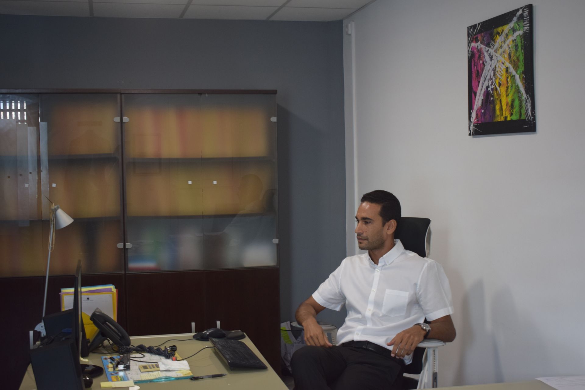 Une personne vêtue d'une chemise blanche boutonnée est assise à un bureau devant une grande armoire en bois sombre.