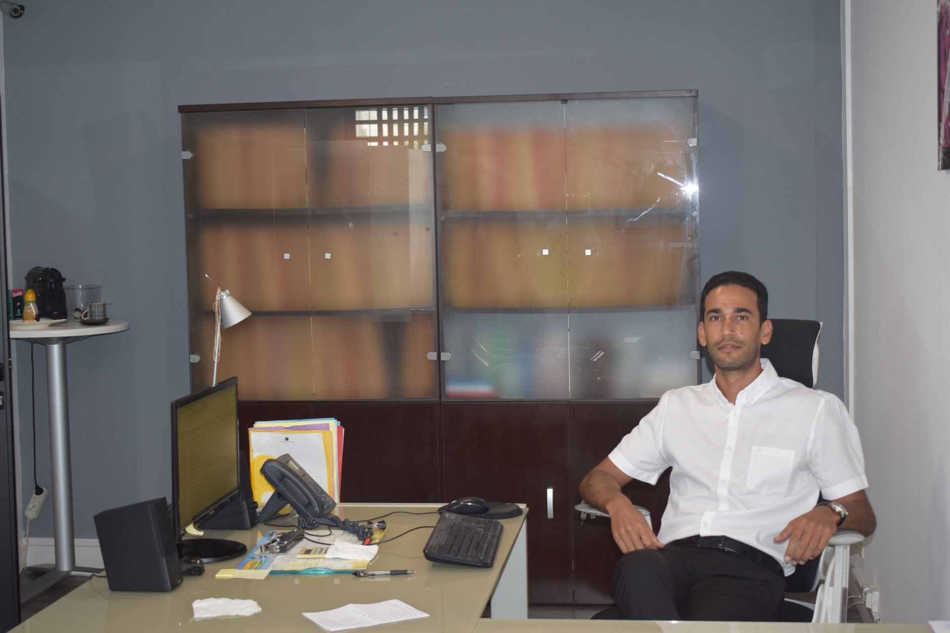 Une personne vêtue d'une chemise blanche à col est assise à un bureau avec un ordinateur et un téléphone, devant une armoire à porte vitrée.