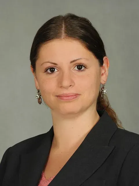 Mujer de cabello castaño con blazer, sonriendo levemente y con aretes. Fondo gris.