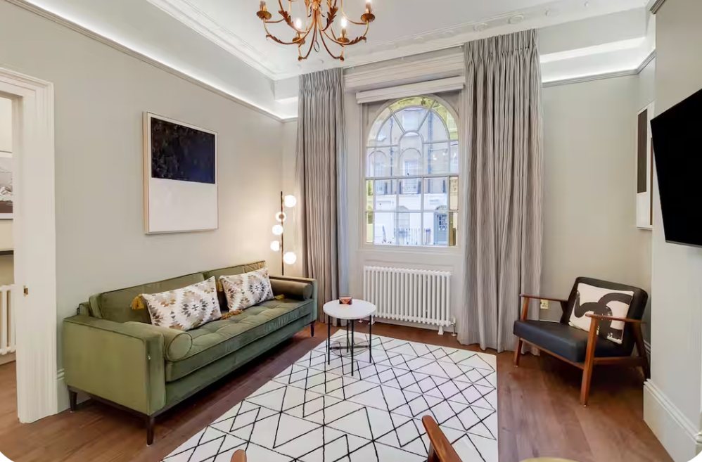 A bright bedroom with a bed featuring patterned cushions and white bedding, wooden floors, two bedside lamps, and art.
