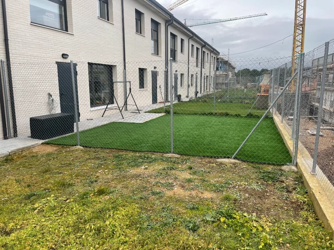 Patio trasero con césped artificial nuevo, vallado entre edificio y obra.