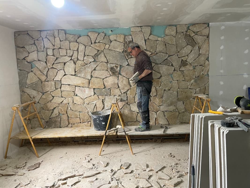 Hombre instalando revestimiento de piedra en una pared, de pie sobre un andamio. Piedras beige y marrones.