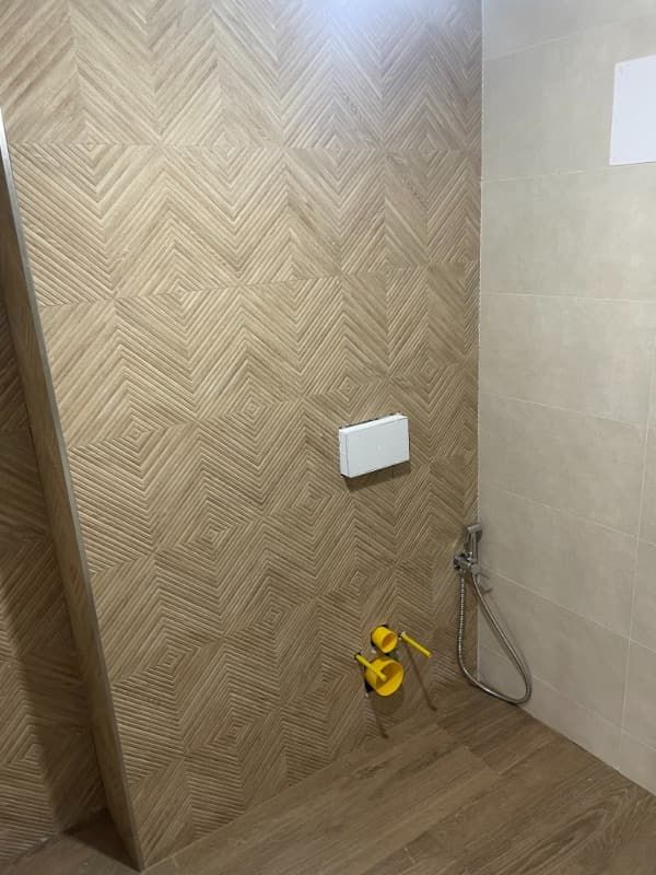 Baño con azulejos de pared texturizados de color beige, piso beige, inodoro con descarga blanca y accesorios de plomería de color amarillo.