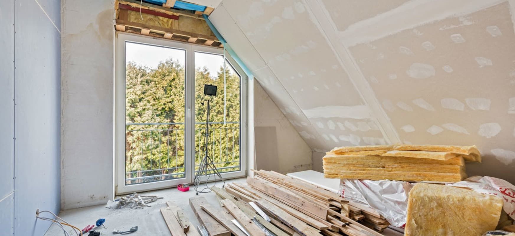 Interior del ático en construcción con ventana, paneles de yeso, aislamiento y madera visibles.