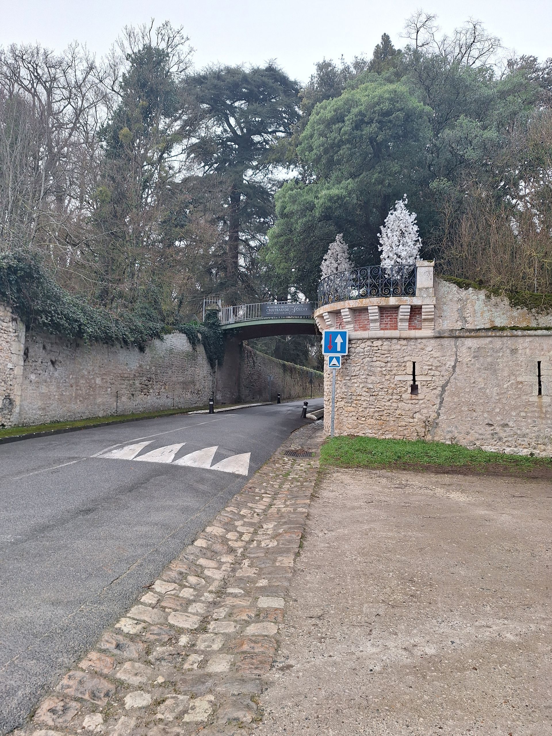 Route menant à un mur de pierre avec un pont au-dessus, des arbres et un ciel nuageux en arrière-plan.