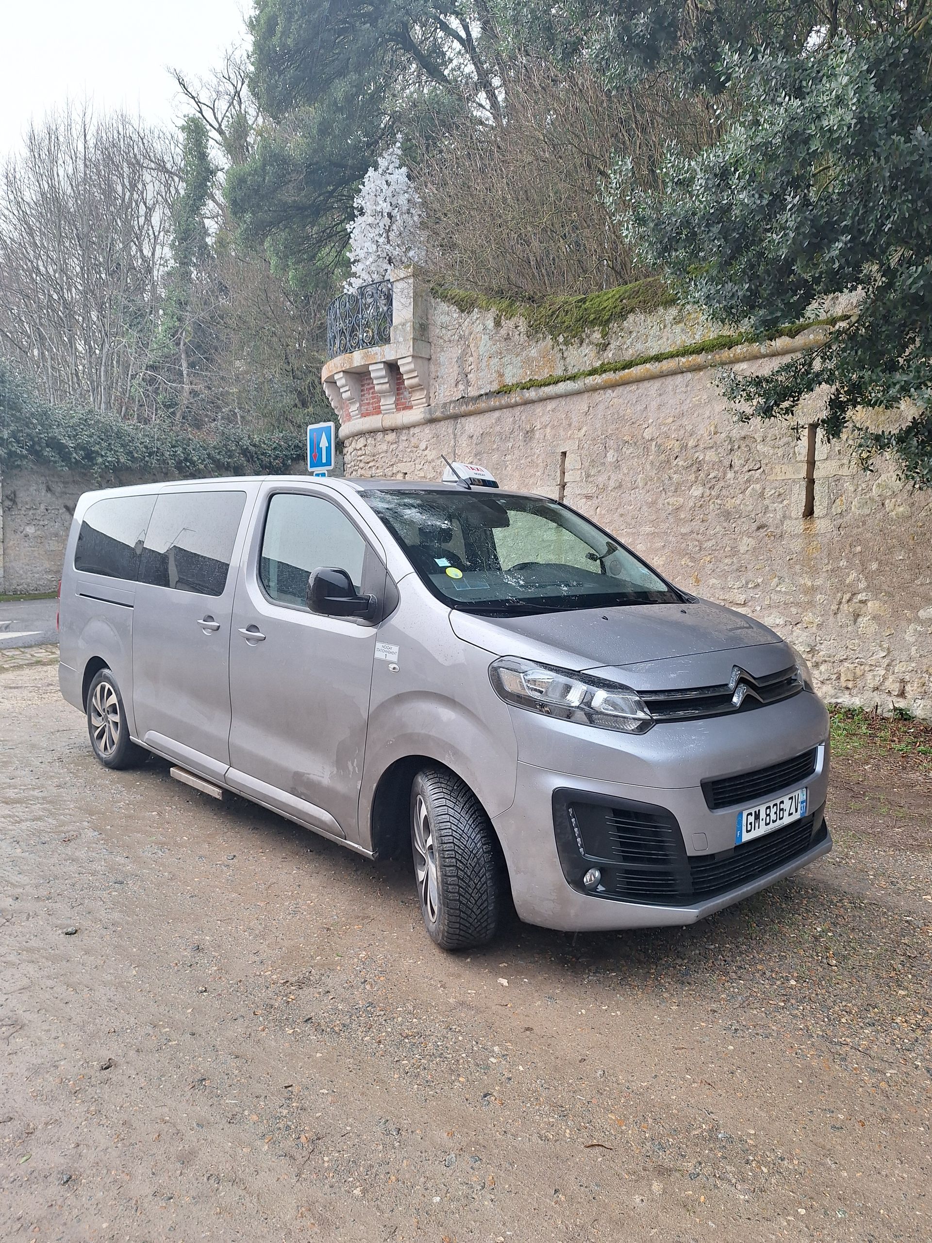 Minivan gris garé sur un chemin de terre à côté d'un muret en pierre ; temps couvert.