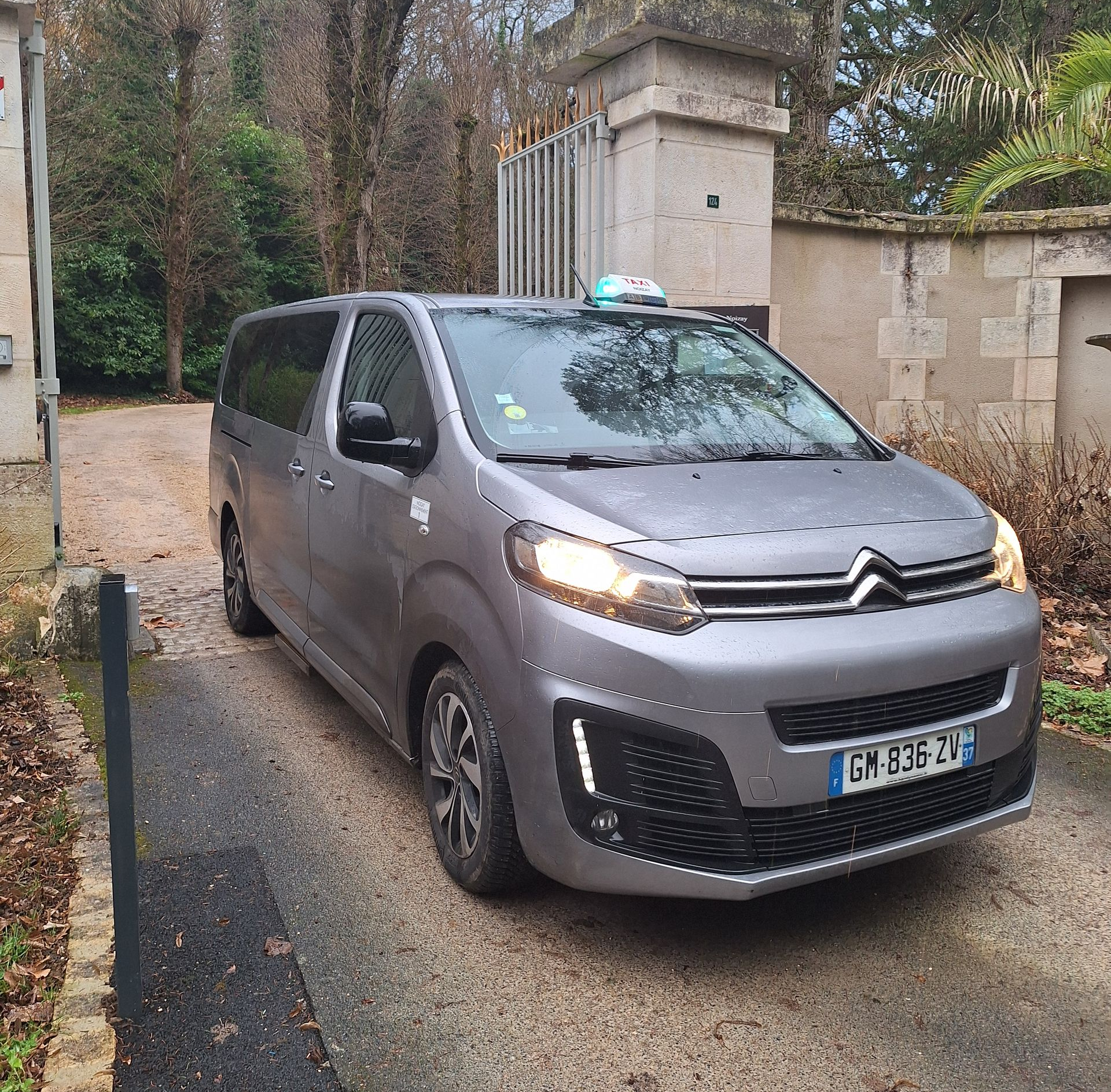 Un taxi gris est stationné dans une allée devant un bâtiment en pierre avec des grilles en fer forgé.