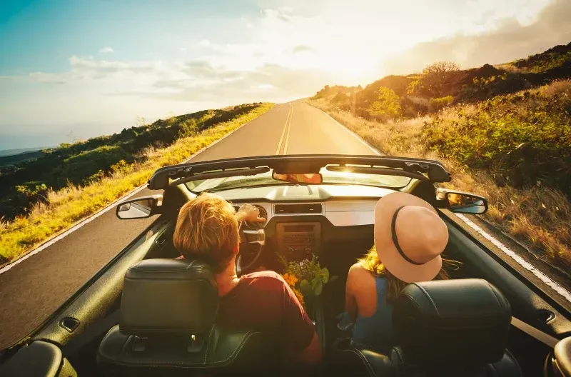 Un hombre y una mujer conducen un automóvil descapotable por una carretera rural.