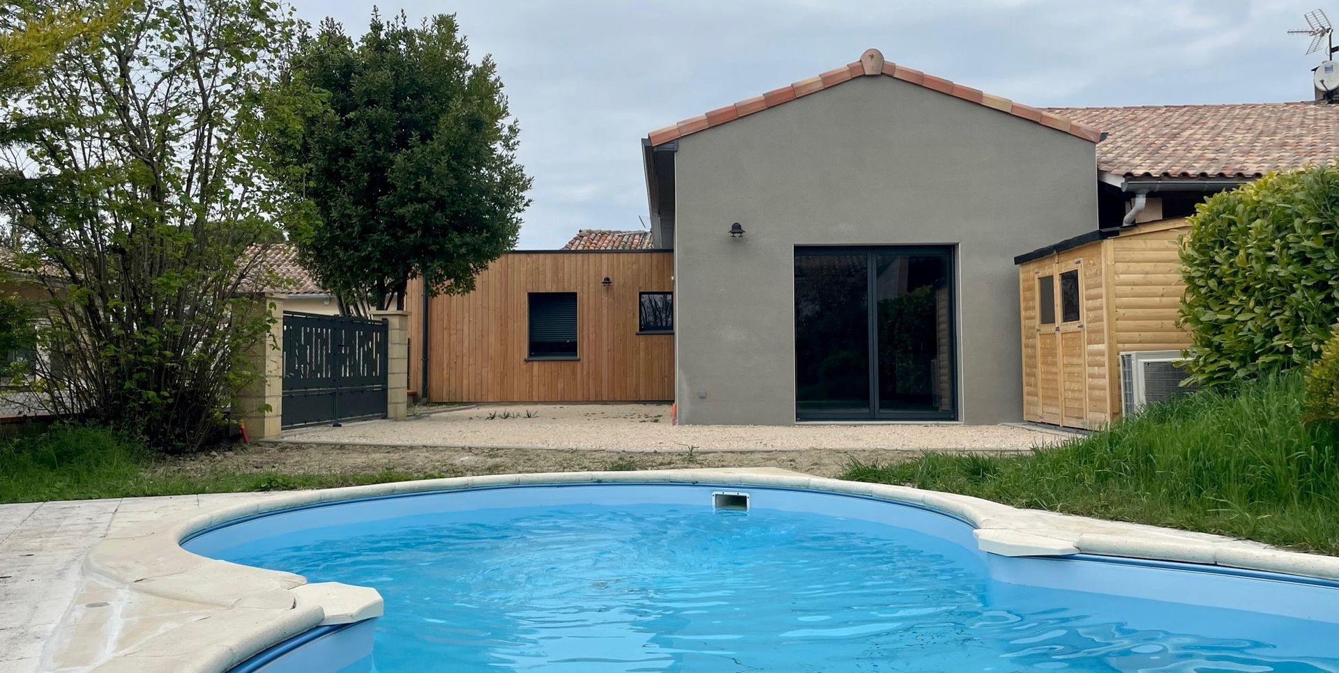 Jardin avec piscine, maison à bardage en bois, arbres et ciel nuageux.