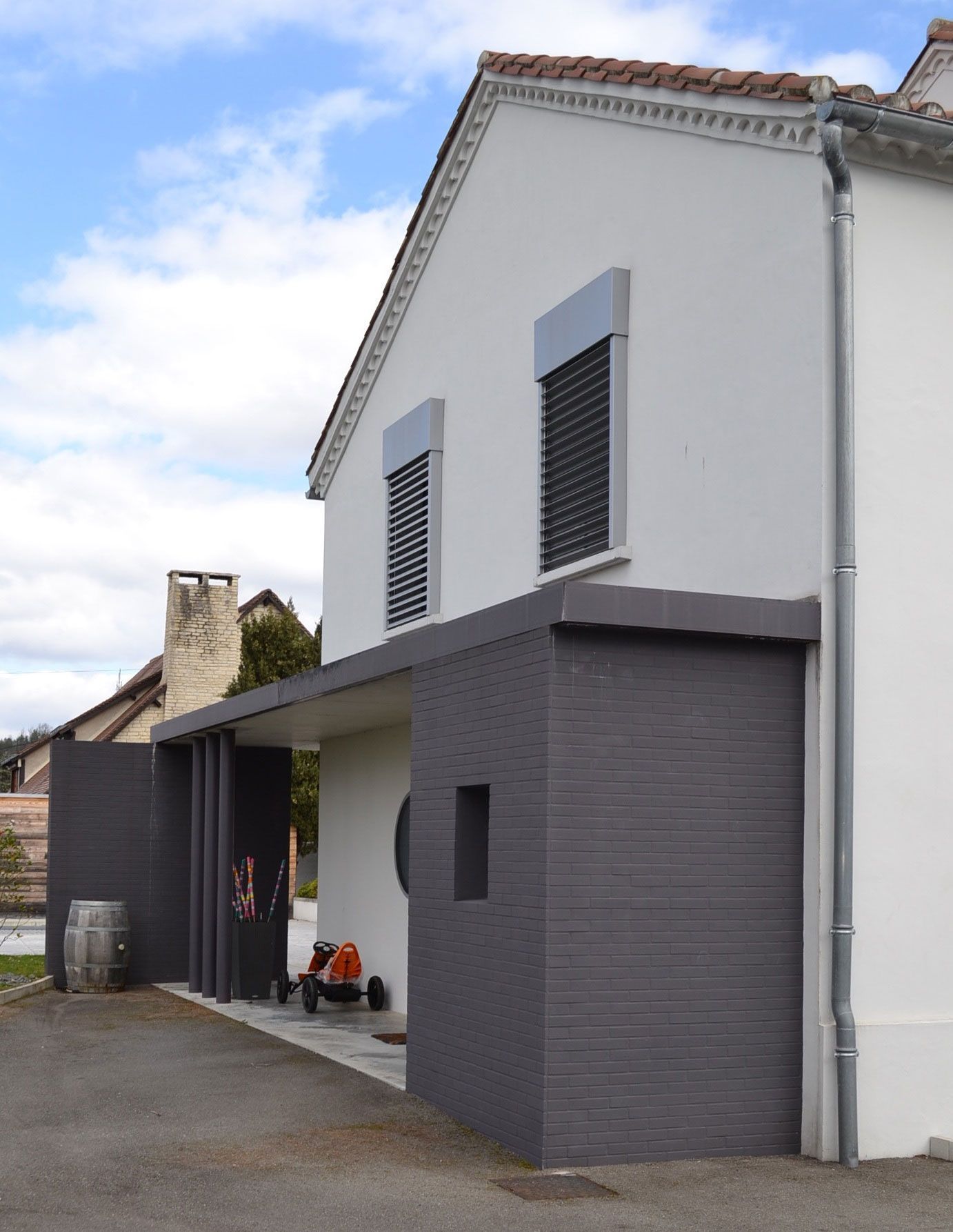 Maison blanche à deux étages avec une extension gris foncé. Les volets et les boiseries sont gris. Un fût et du matériel sont visibles.