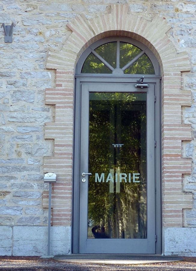 Porte grise avec fenêtre cintrée en verre ; inscription MAIRIE sur le verre ; située dans un bâtiment en pierre avec des accents de brique.