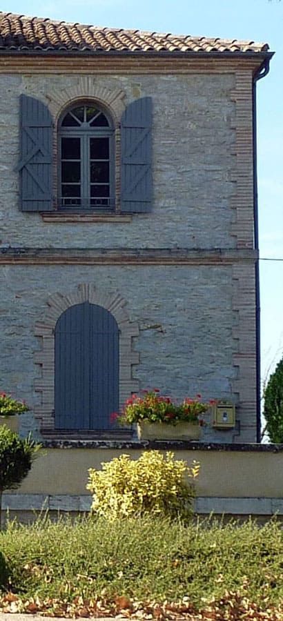 Bâtiment de deux étages avec des volets bleus et un toit de tuiles, entouré de verdure et d'un ciel bleu.