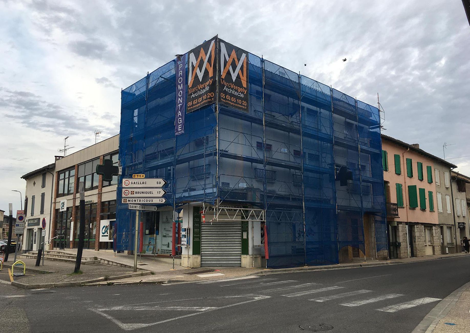 Bâtiment en rénovation, recouvert d'un filet bleu, à un coin de rue, sous un ciel nuageux.