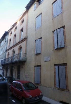 Vue de rue d'un immeuble de grande hauteur aux volets gris et d'une voiture rouge garée devant.