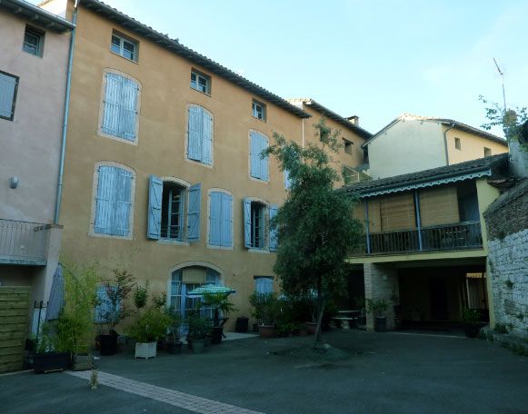 Bâtiment jaune de trois étages avec volets bleus, cour et petit arbre.