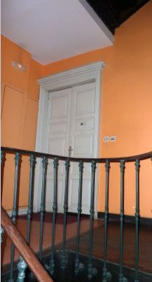 Couloir aux murs orange avec rampe d'escalier en bois foncé, menant à une double porte blanche.